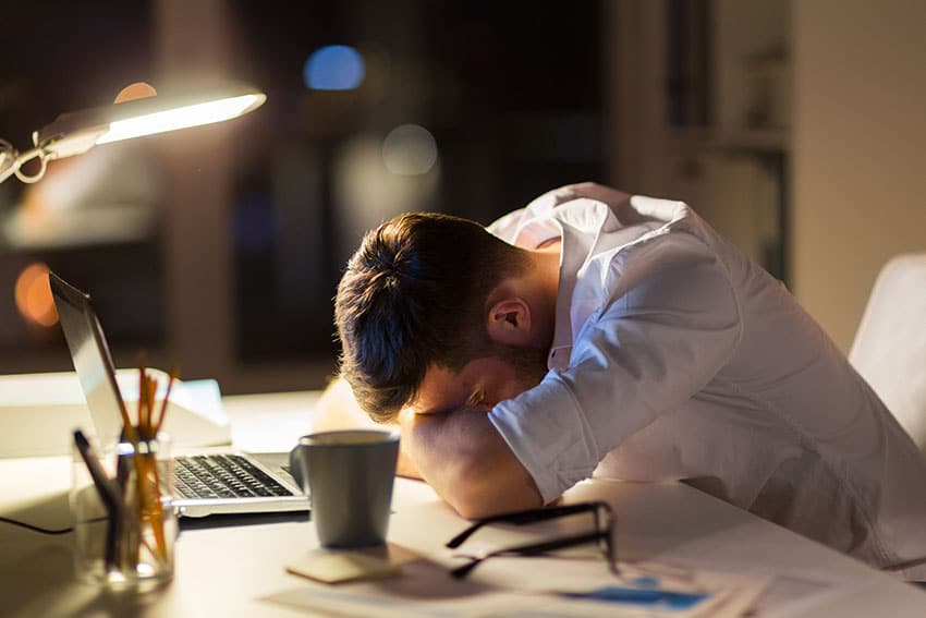 exhausted man who fell asleep at his computer with single light on and empty cup of coffee